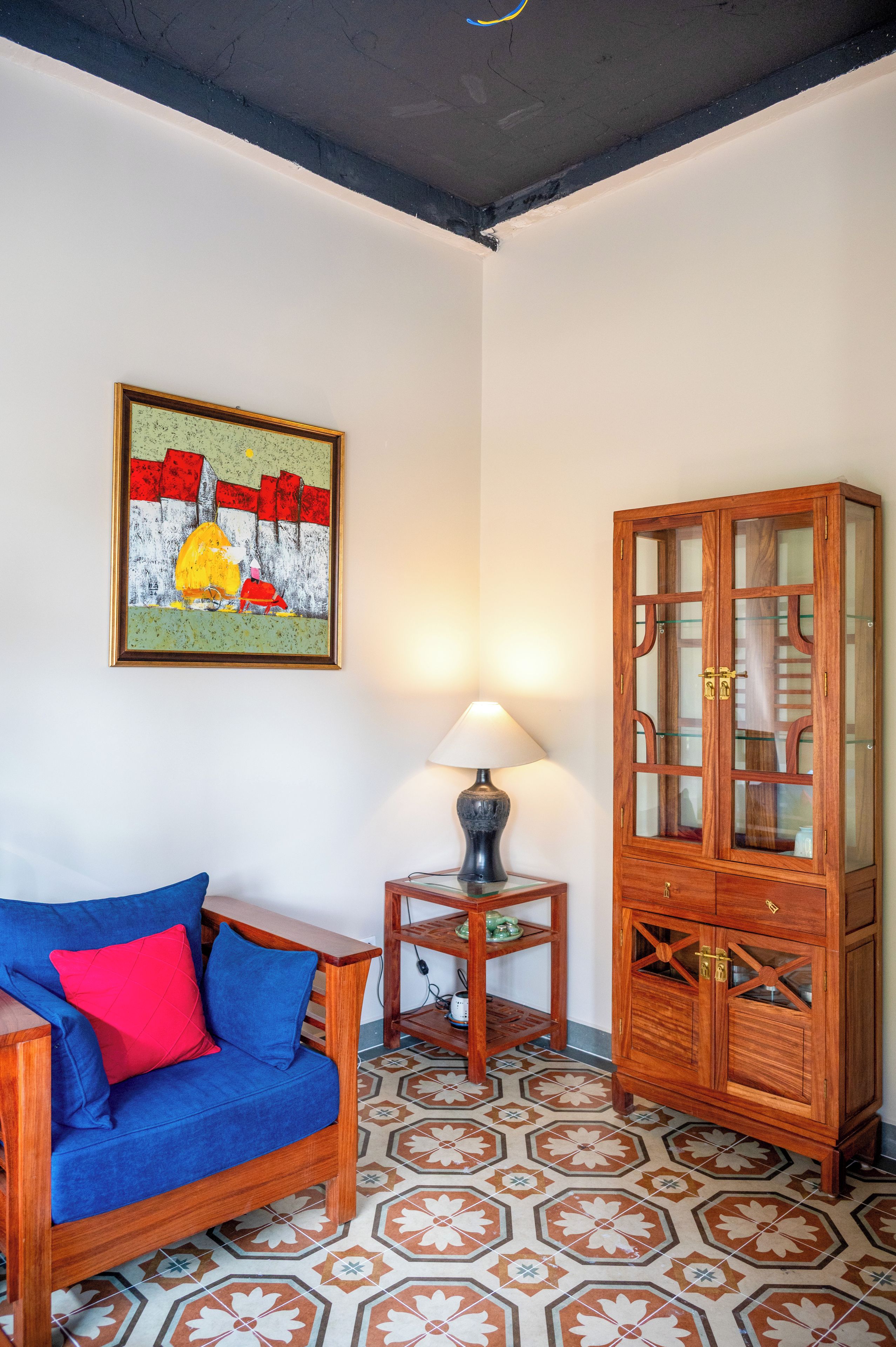 Una habitación con un sofá azul, un mueble de madera con puertas de cristal, una mesa con lámpara y un suelo estampado.