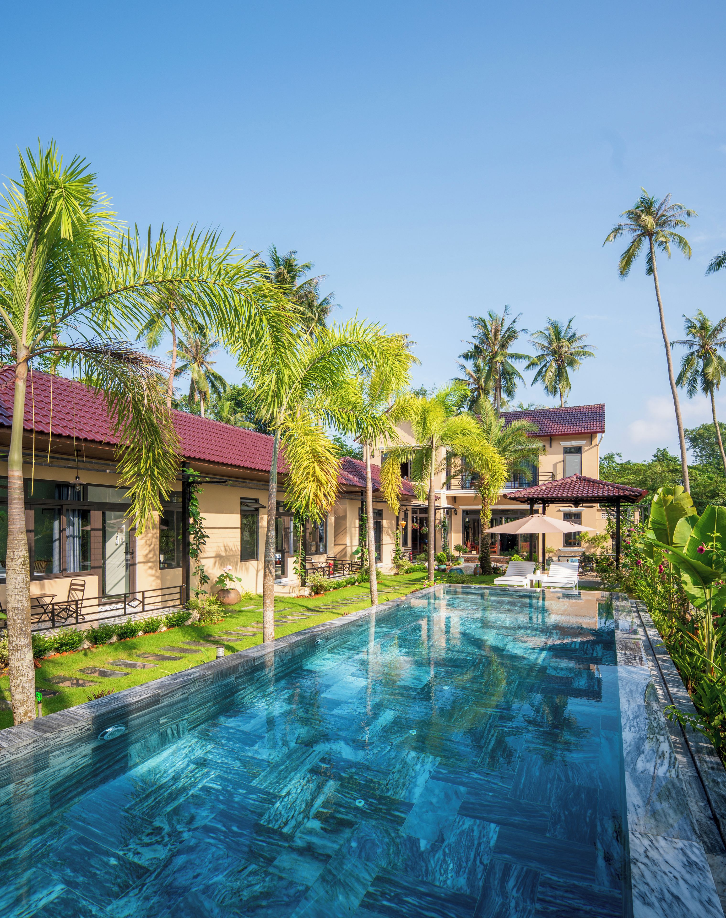 A swimming pool with a clear blue water surface, surrounded by palm trees and a building with a red-tiled roof.