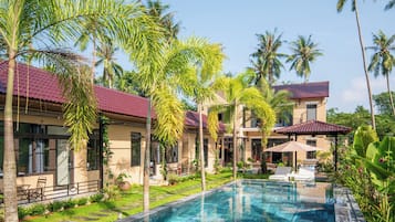 A swimming pool with a clear blue water surface, surrounded by palm trees and a building with a red-tiled roof.