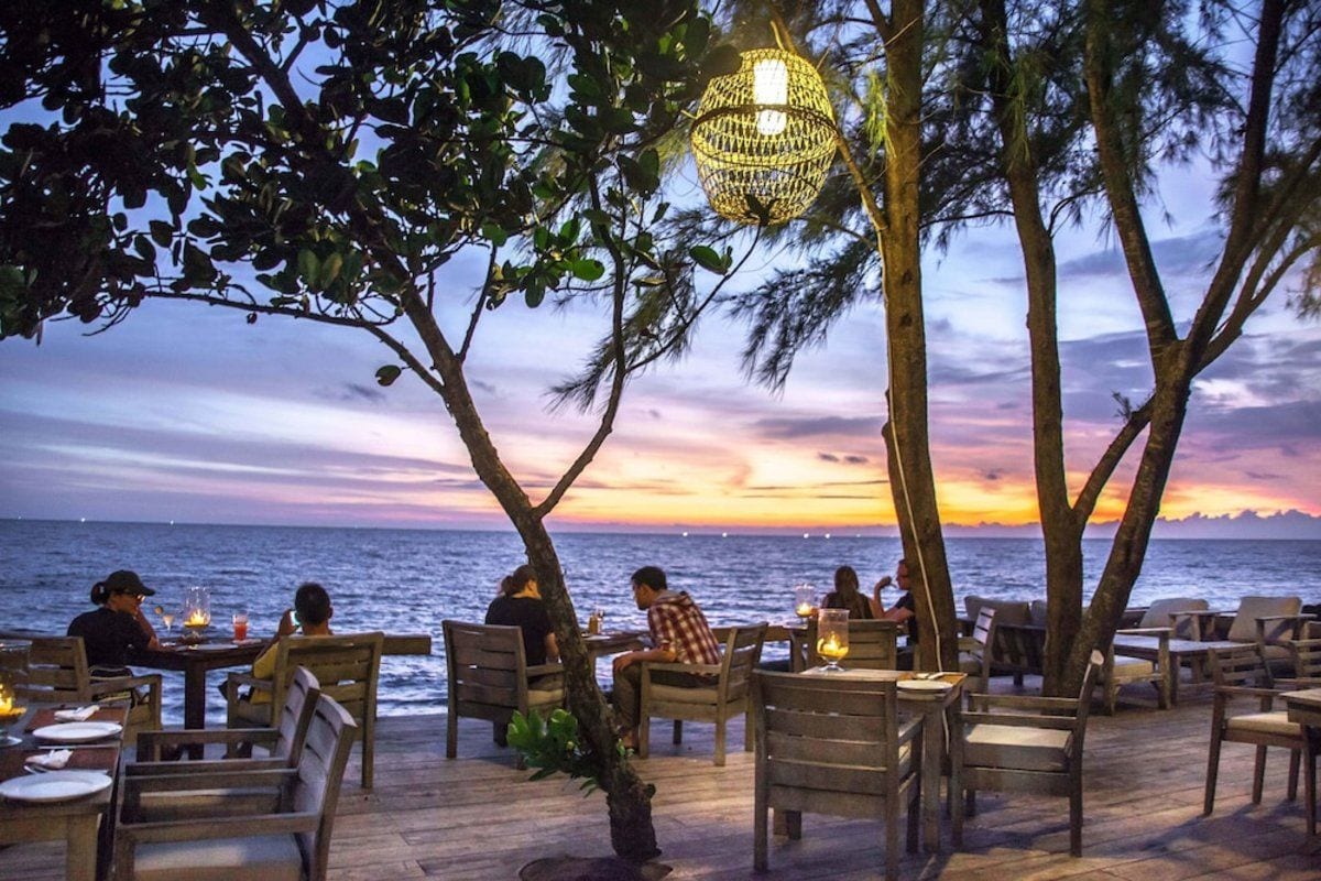 Un espacio de comedor al aire libre con mesas y sillas, con vista al mar, y un atardecer de fondo.