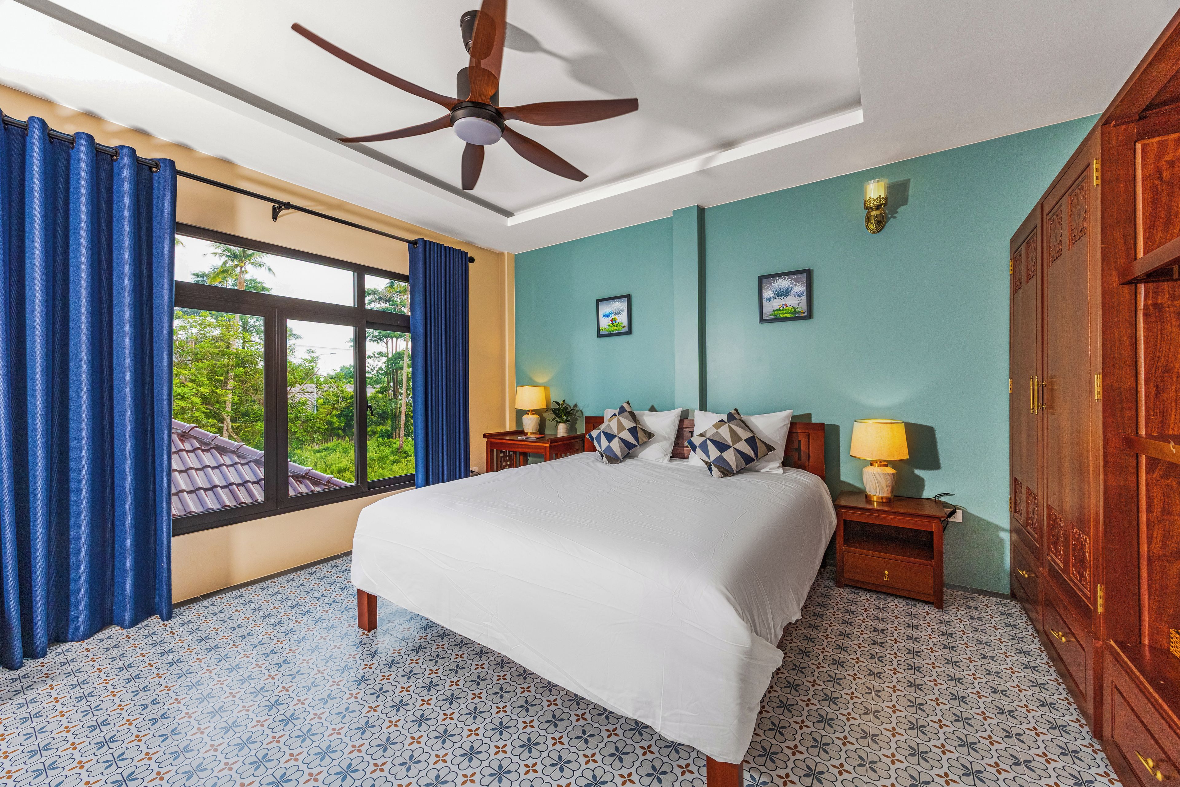A hotel room with a bed, two bedside tables, a ceiling fan, a window with curtains, and a view of greenery outside.