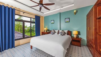 A hotel room with a bed, two bedside tables, a ceiling fan, a window with curtains, and a view of greenery outside.