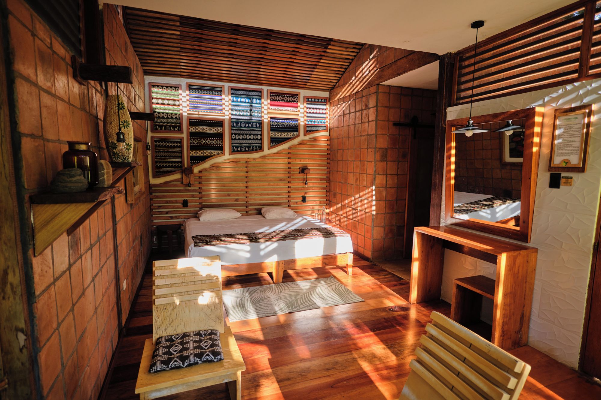 Ein gemütliches Zimmer mit einem Bett, einem kleinen Tisch und einer Bank. Es gibt ein Fenster mit gemusterten Vorhängen und eine Holzbodenverkleidung.
