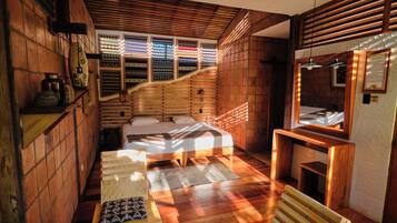 Ein gemütliches Zimmer mit einem Bett, einem kleinen Tisch und einer Bank. Es gibt ein Fenster mit gemusterten Vorhängen und eine Holzbodenverkleidung.