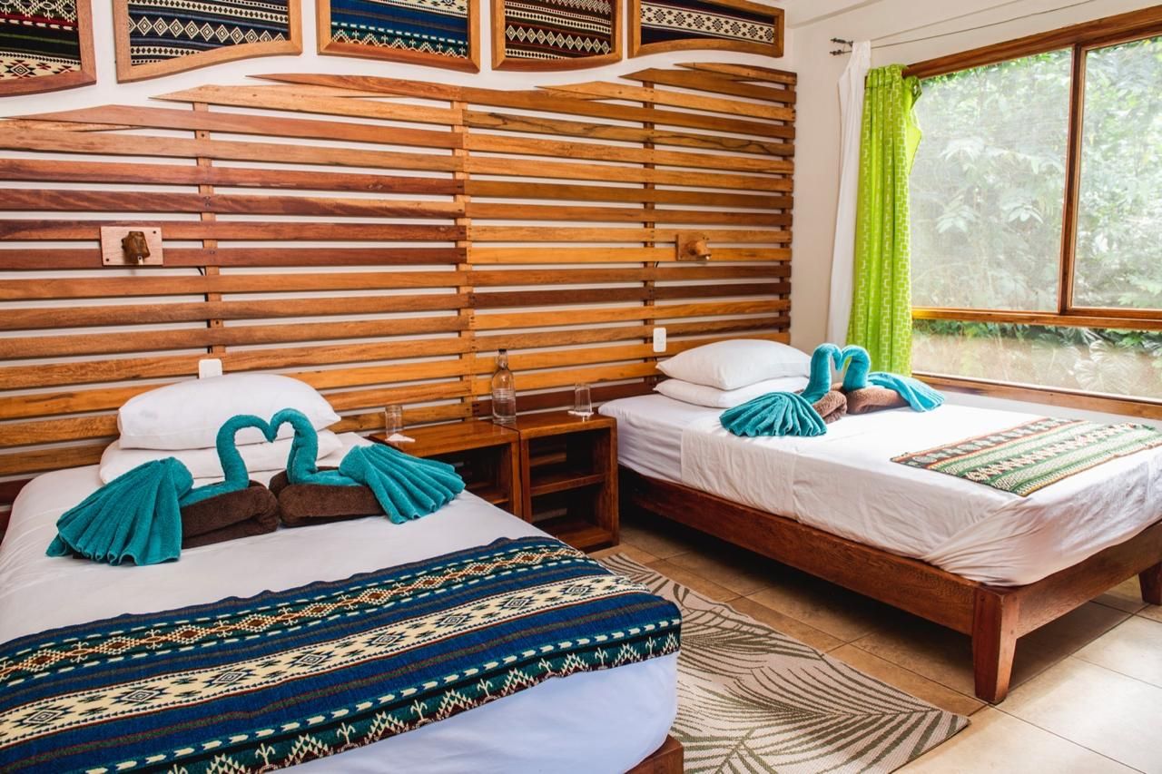 Ein Zimmer mit zwei Holzbett, einem Holzkopfstück, einem Fenster mit Vorhängen und einem gemusterten Teppich.