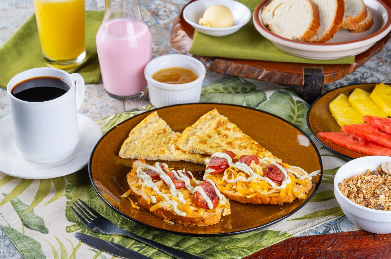 Ein Frühstückstisch mit Toast, belegt mit Käse und Wurst, ein Glas Orangensaft, eine Tasse Kaffee und eine Schüssel Granola.