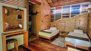 A room with two beds, a wooden bench, a mirror, and a wall with patterned textiles.
