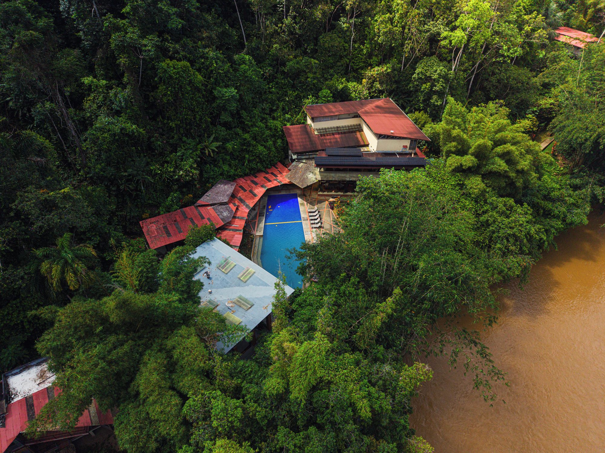 Ein abgelegenes Resort mit Swimmingpool, umgeben von dichtem Wald, einem kleinen Gebäude mit rotem Dach und einem Fluss in der Nähe.