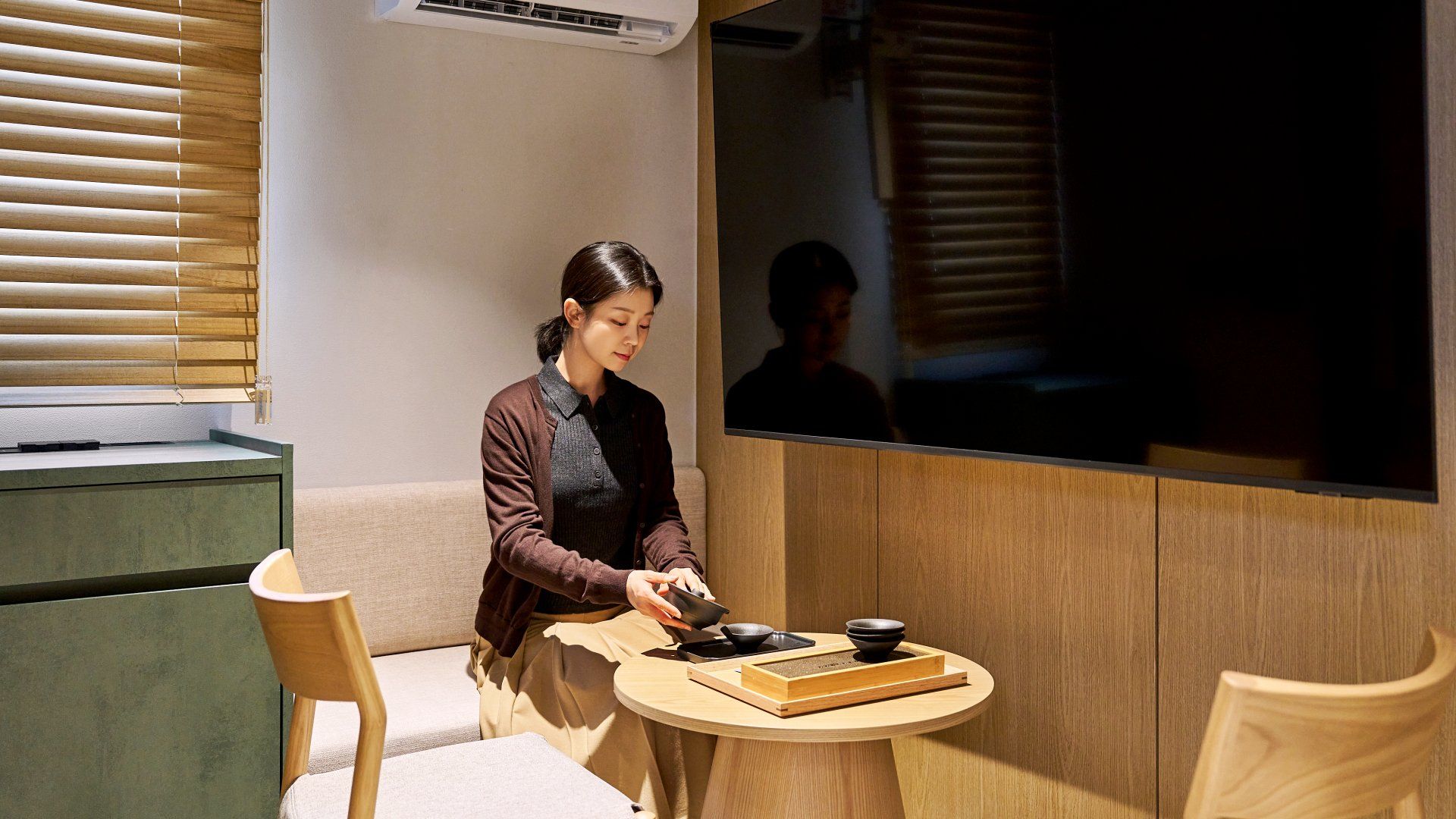 A person seated at a round wooden table with a bowl and chopsticks, in a room with a large window and a wall-mounted TV.