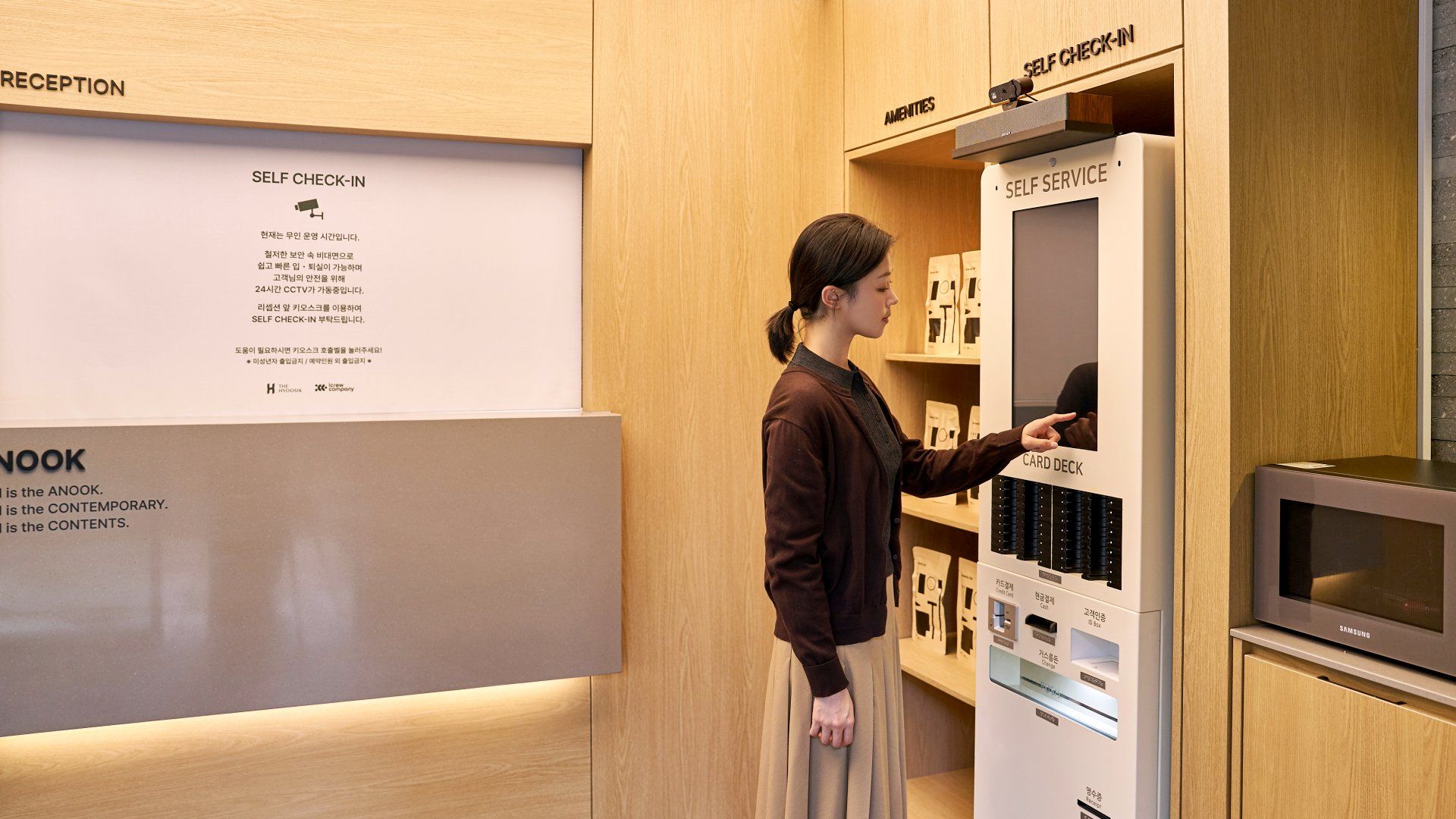 Una persona utilizando un quiosco de autoservicio en un hotel, con paneles de madera y secciones identificadas para recepción, servicios y autoservicio.