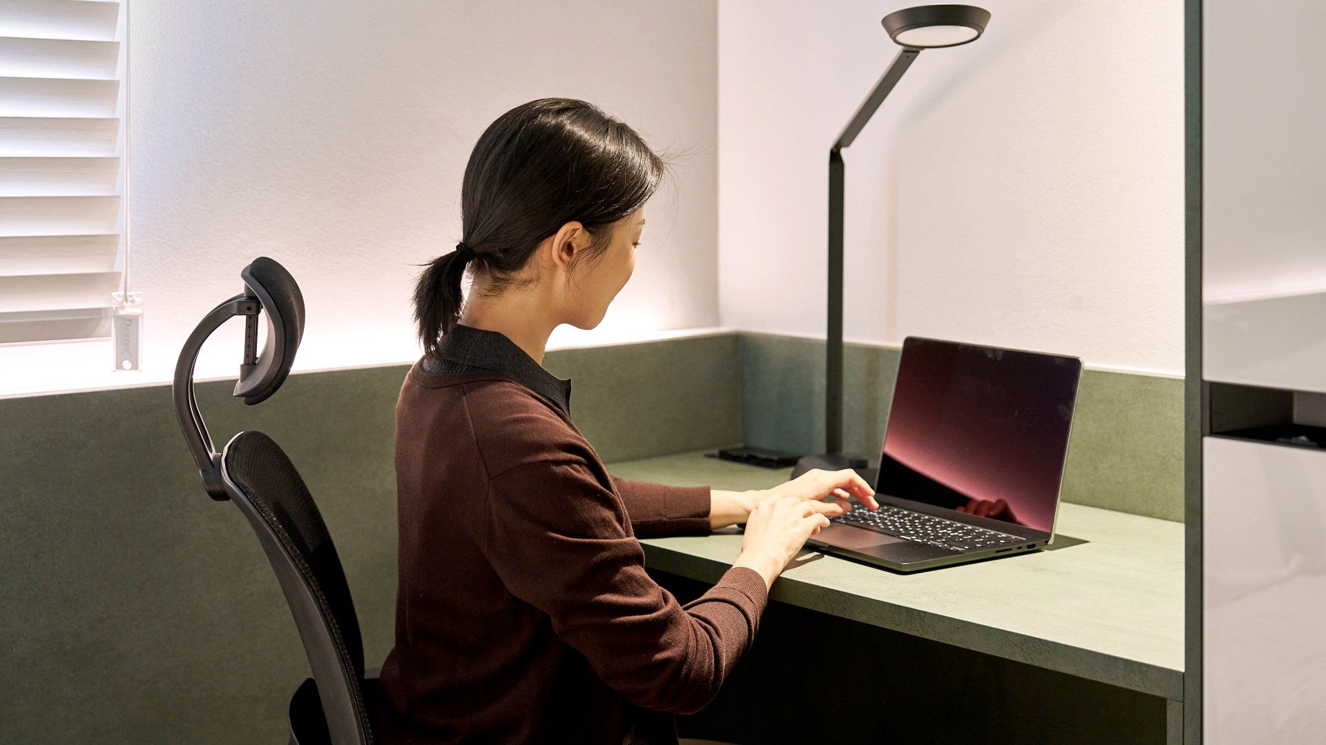 Una persona sentada en un escritorio con una computadora portátil, una silla y una lámpara de escritorio en una habitación con una ventana y una pared.