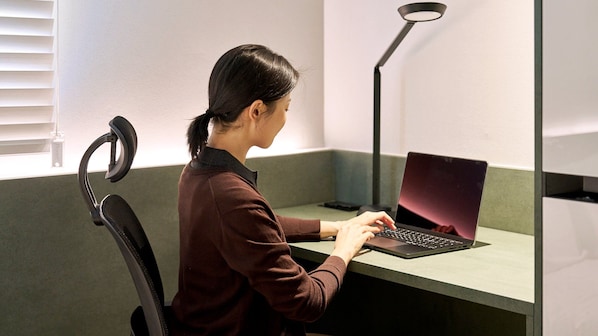 A person seated at a desk with a laptop, a chair, and a desk lamp in a room with a window and a wall.