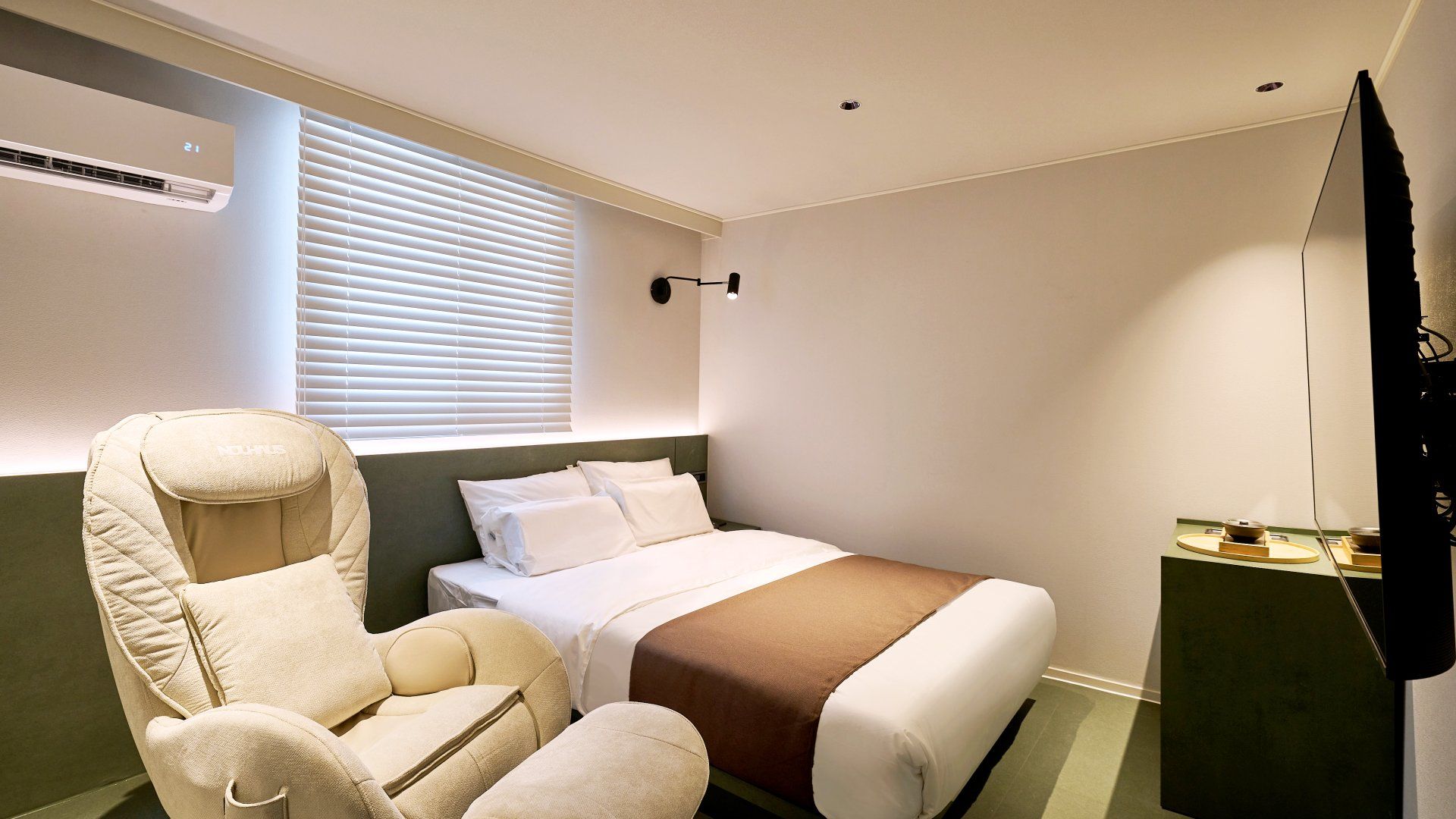 A hotel room with a bed, a chair, a TV, and a wall-mounted air conditioner.