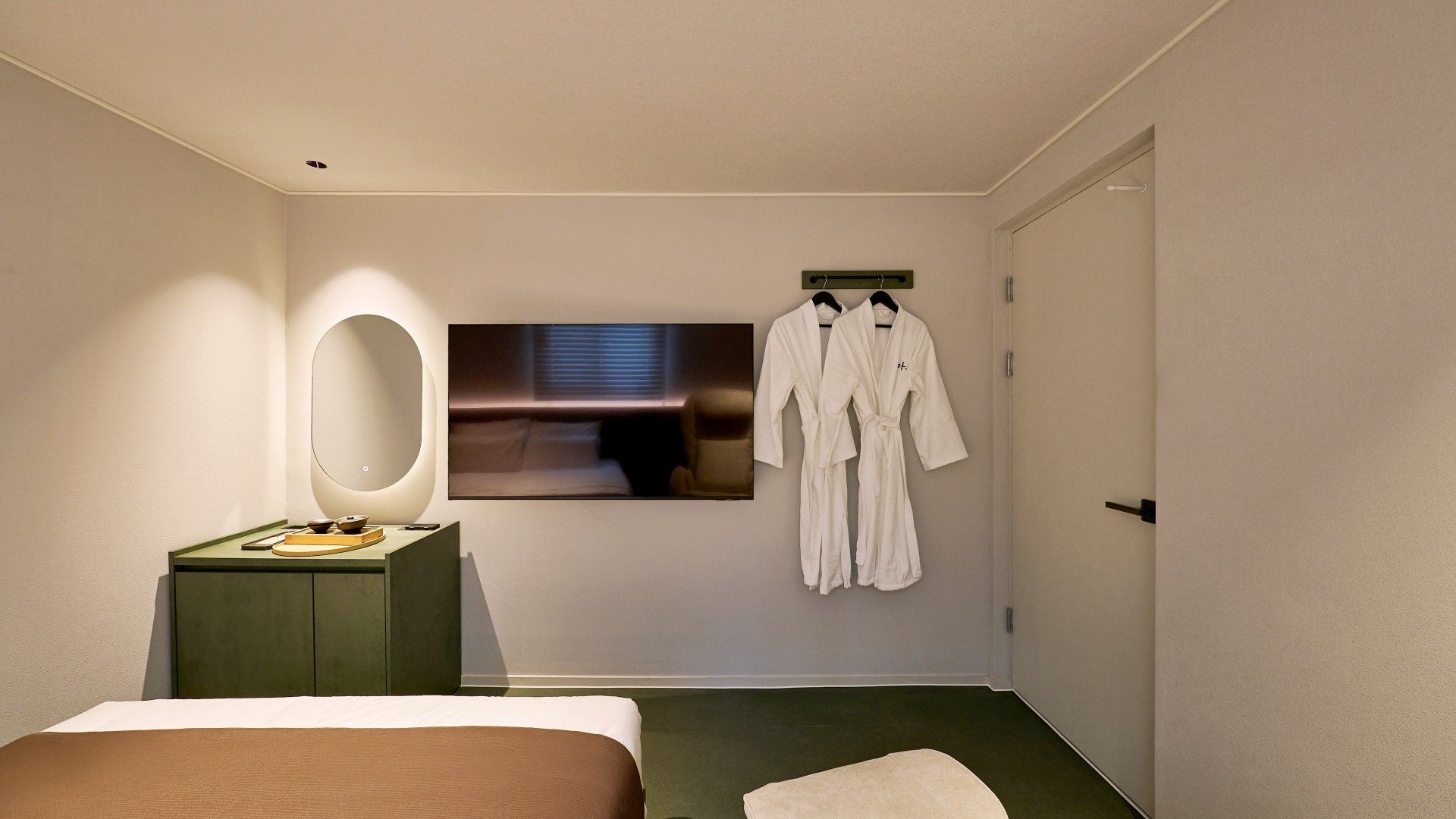 Habitación de hotel con cama, mesita de noche con lámpara, espejo, televisor de pared y bata colgada de un perchero.
