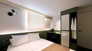 A hotel room with a bed, bedside table, lamp, wardrobe, and a window with blinds.