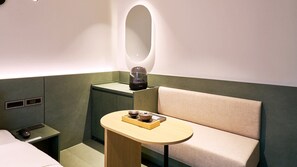 Habitación de hotel compacta con cama, mesita de noche pequeña, una mesa redonda con bandeja, sofá y espejo montado en la pared.