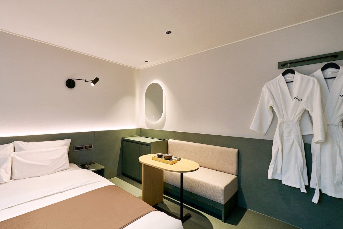 A hotel room with a bed, a bedside table with a bowl and a book, a small round table with a bowl, a robe rack with two robes, and a wall-mounted light fixture.
