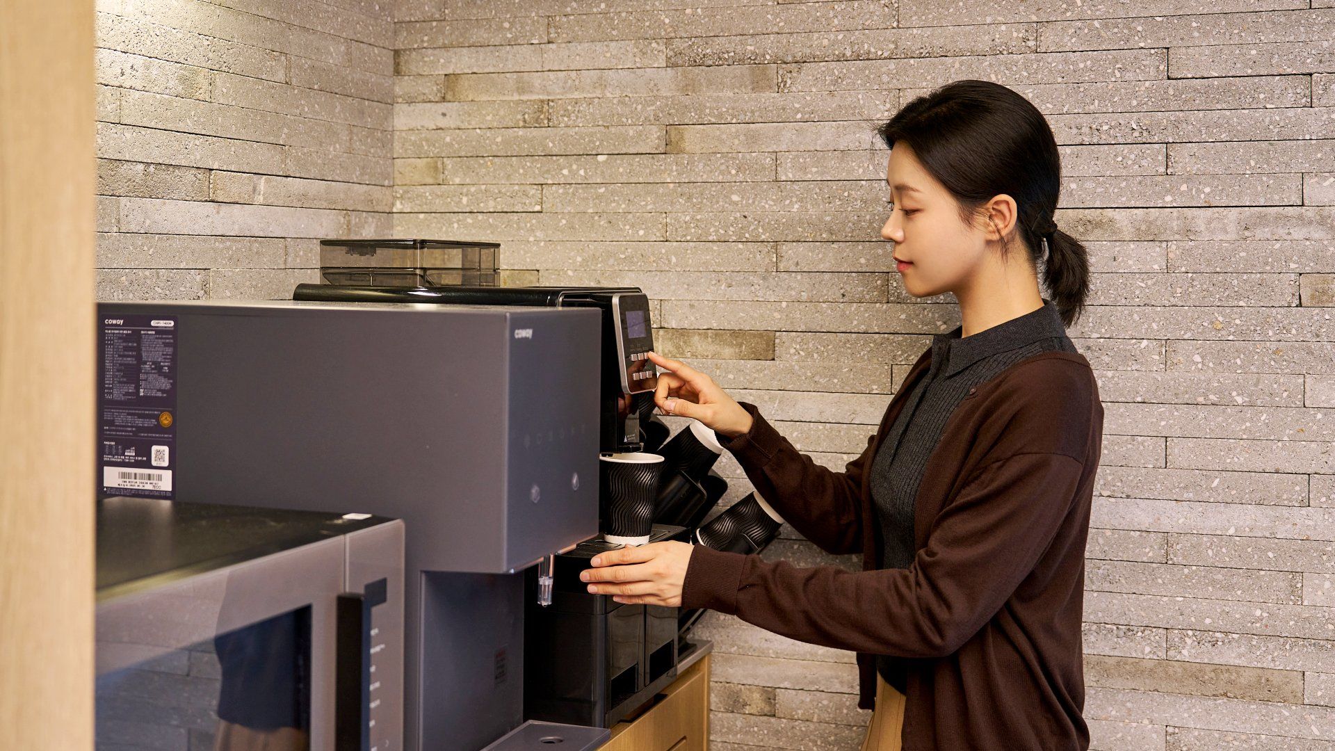Una persona operando una máquina de café con una interfaz digital, en una habitación con pared de ladrillo.