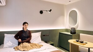 A modern hotel room with a bed, a bedside table with a kettle, a small table with a teapot, and a wall-mounted lamp.