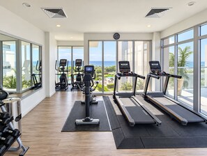 Ein Fitnessstudio mit Laufbändern, Crosstrainern und Kurzhanteln, große Fenster mit Blick auf das Meer.