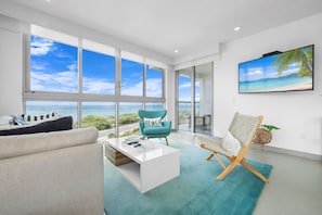 Ein modernes Wohnzimmer mit einem großen Fenster mit Blick auf einen Strand, einem weißen Couchtisch, einem türkisfarbenen Sessel und einem Korbstuhl.
