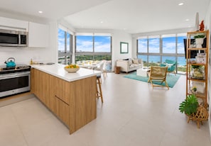 A modern kitchen with a large island, a dining area with a view, and a living space with a sofa and chairs. - Coastal Elegance I Eagle Beach Ocean View I 4th Fl (Oranjestad)