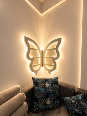 Ein Zimmer mit einer wandmontierten Lampe in Form eines Schmetterlings, einem dunkelgrauen Sofa mit gemusterten Kissen und einer Ecke mit einer hellen Wand.