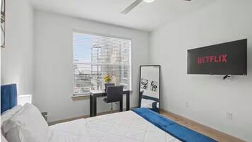 Habitación de hotel con cama, escritorio, silla, ventana con persianas, ventilador de techo y televisor montado en la pared.
