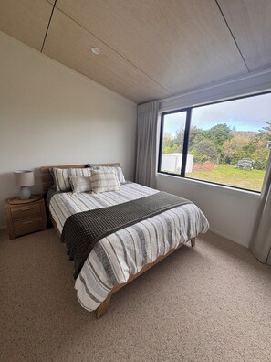 A bedroom with a large bed, a nightstand, a lamp, and a window with a view of greenery. - WAINUI SUNSET HIDEAWAY
WHERE EVERY EVENING ENDS WITH A BEAUTIFUL SUNSET (Gisborne)