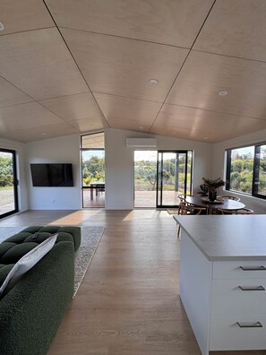 A spacious living area with a large flat-screen TV, a green sofa, a dining table with chairs, and a kitchen island with drawers.