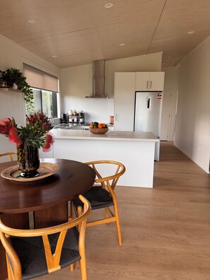 A modern kitchen with a central island, wooden chairs, and a dining table with a vase of flowers. - WAINUI SUNSET HIDEAWAY
WHERE EVERY EVENING ENDS WITH A BEAUTIFUL SUNSET (Gisborne)