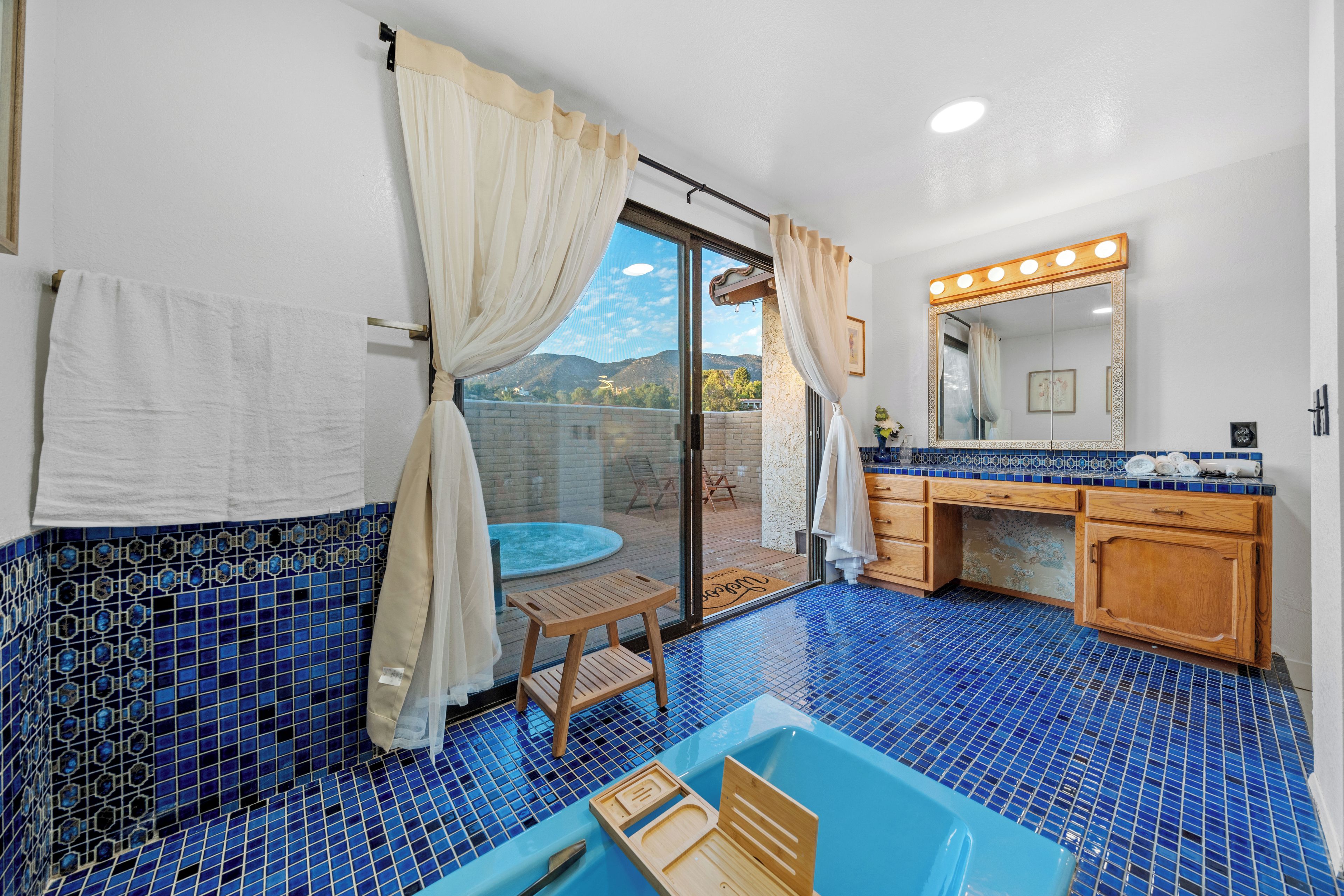 Un baño con piso de azulejos azules, un taburete de madera, una bañera y un tocador con espejo e iluminación.