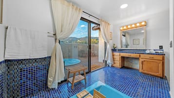 Un baño con piso de azulejos azules, un taburete de madera, una bañera y un tocador con espejo e iluminación.