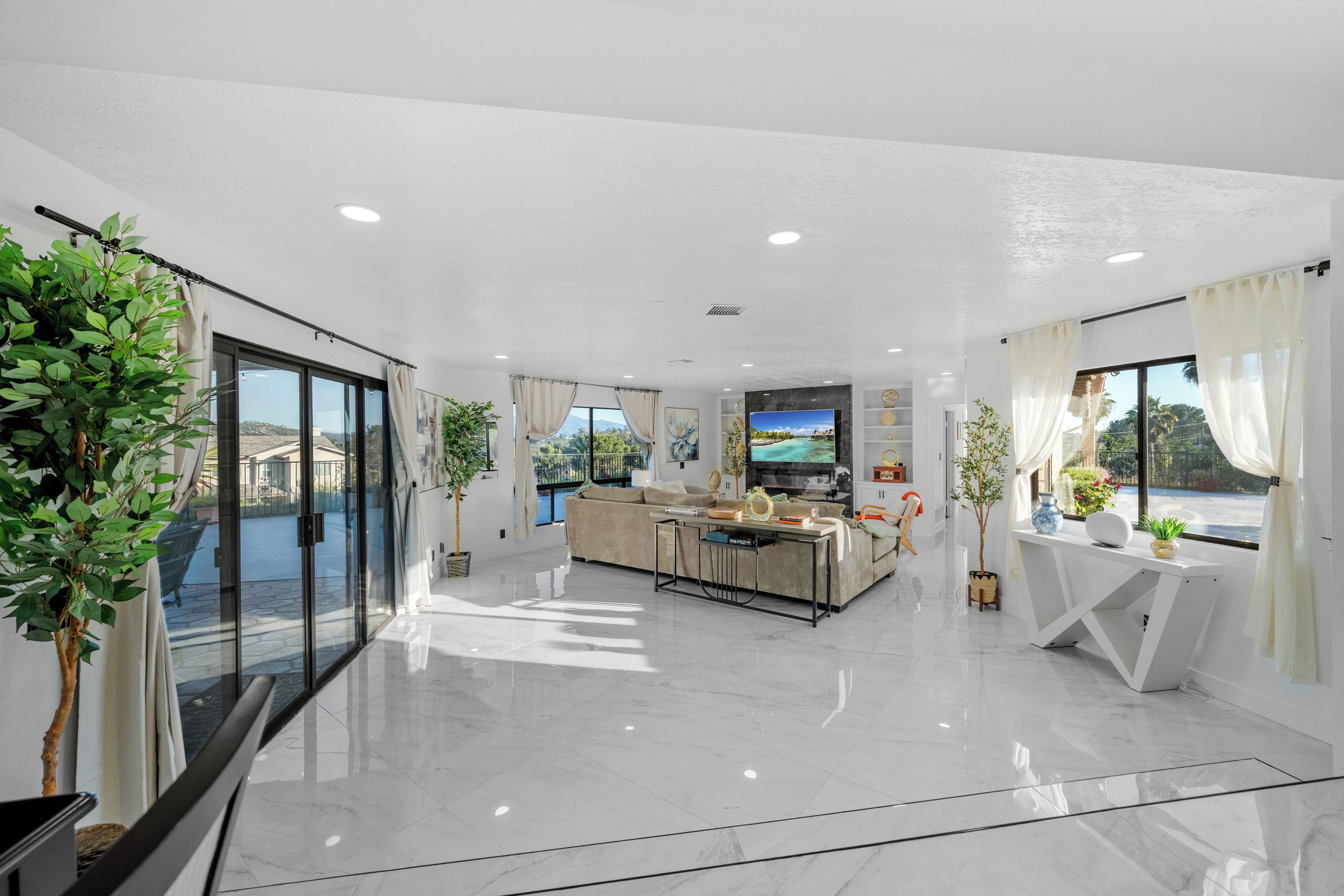 A spacious living area with a sofa, a coffee table, and a TV. There are potted plants and a view of the outdoors.