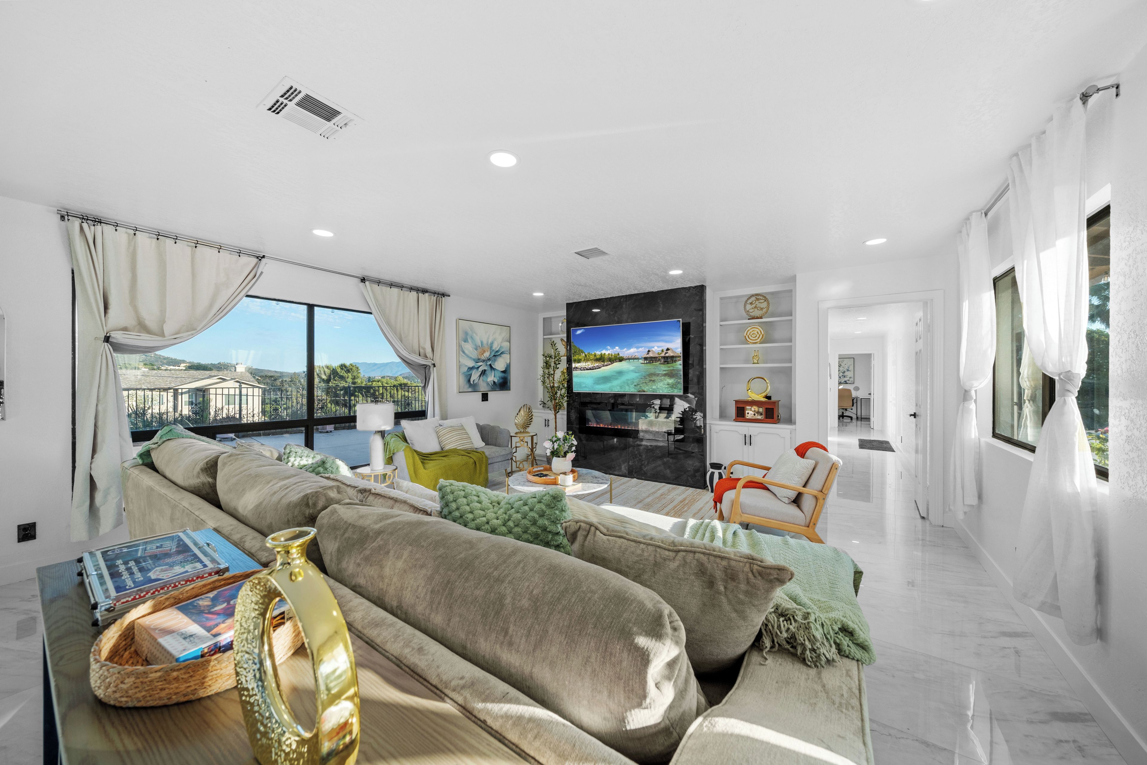 A modern living room with a large sofa, a coffee table with books and a vase, a flat-screen TV, and a view of a building through large windows.