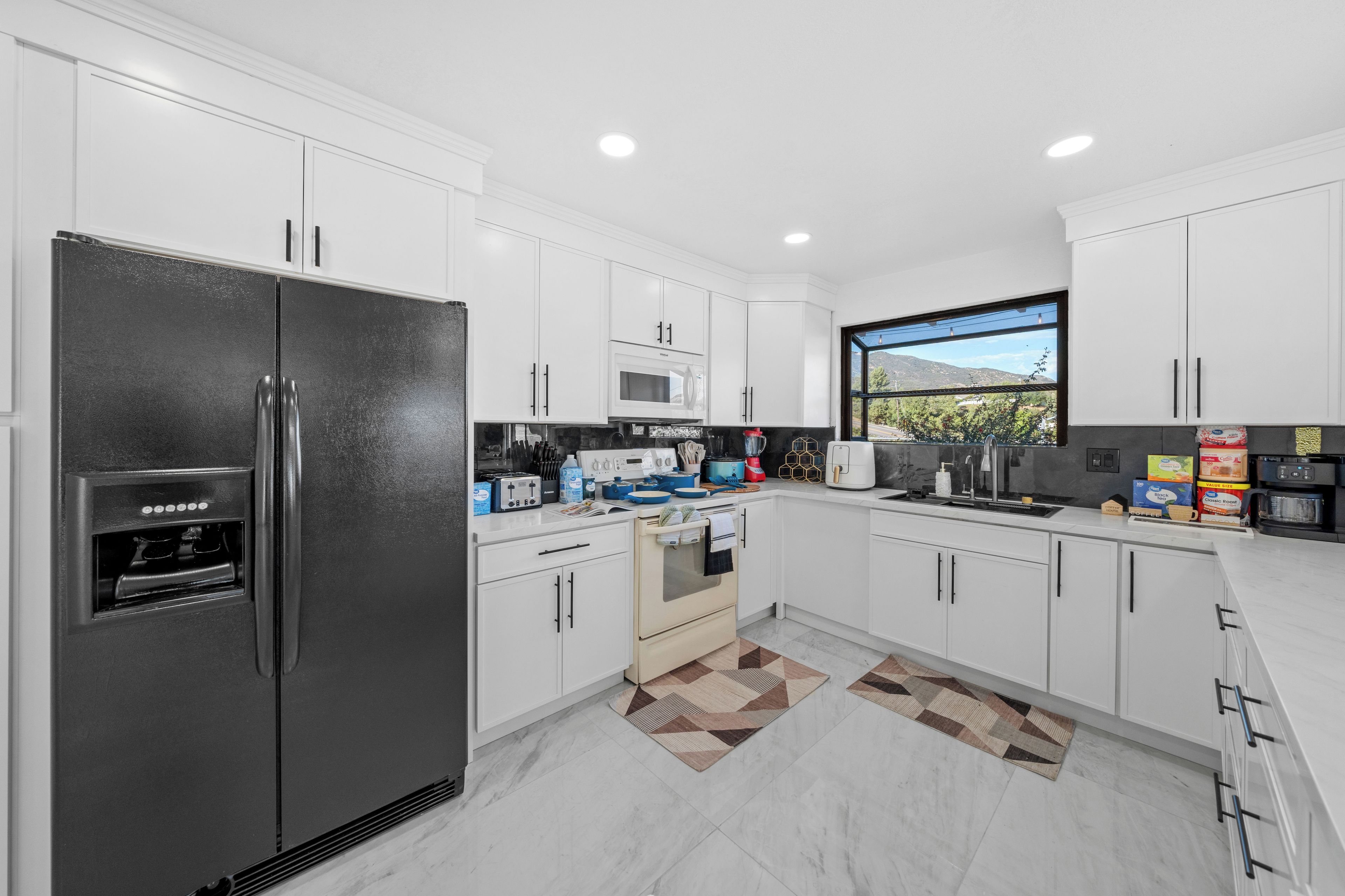 A modern kitchen with white cabinets, a black refrigerator, a microwave, and a window with a view of greenery.