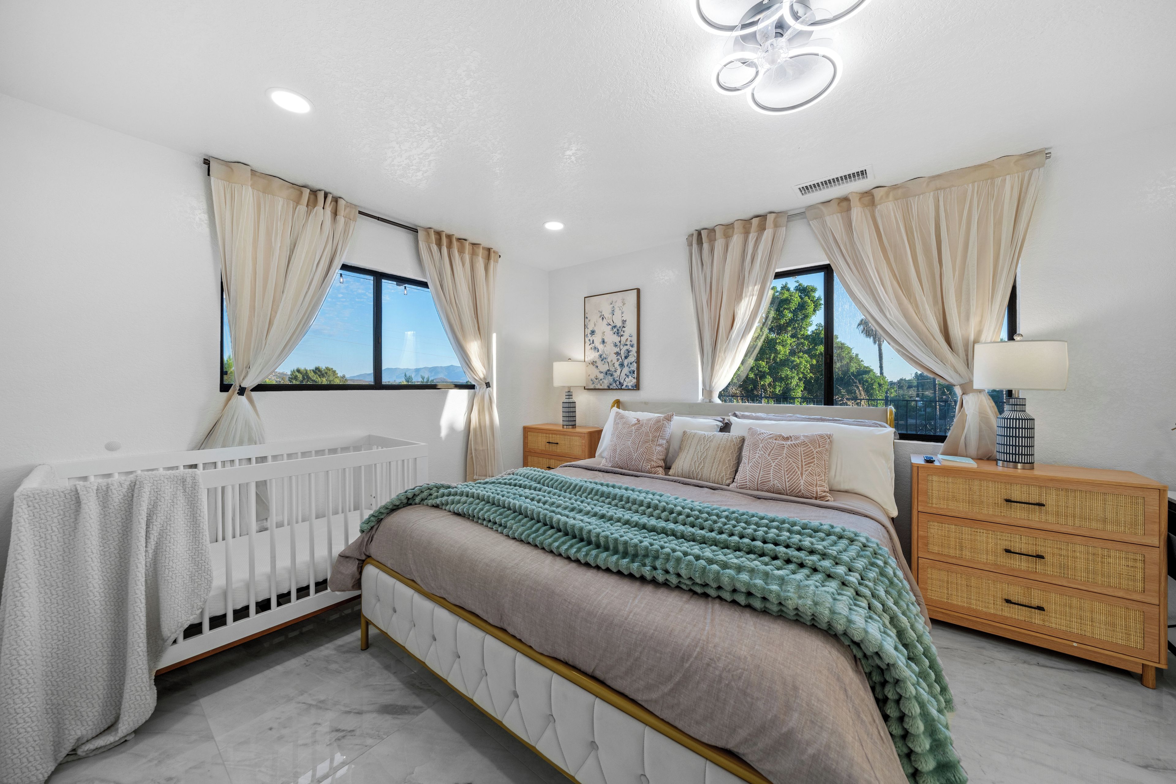 Ein Schlafzimmer mit einem weißen Babybett, einem großen Bett mit grüner Strickdecke, zwei Nachttischen mit Lampen und Blick ins Freie durch die Fenster.