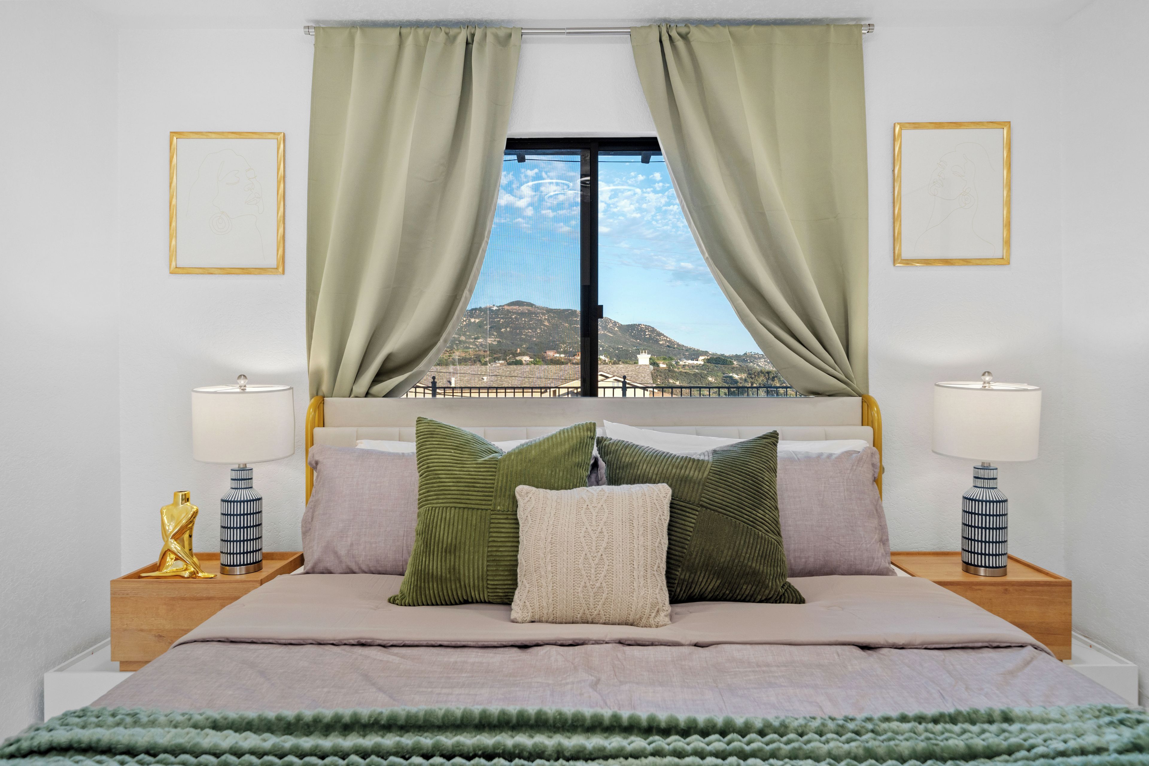 Ein Schlafzimmer mit einem Bett, grünen und beigen Kissen, Blick auf eine Landschaft durch ein Fenster und zwei Nachttische mit Lampen.