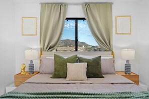 Une chambre à coucher avec un lit, des coussins verts et beiges, une vue sur un paysage par la fenêtre, et deux tables de chevet avec des lampes.