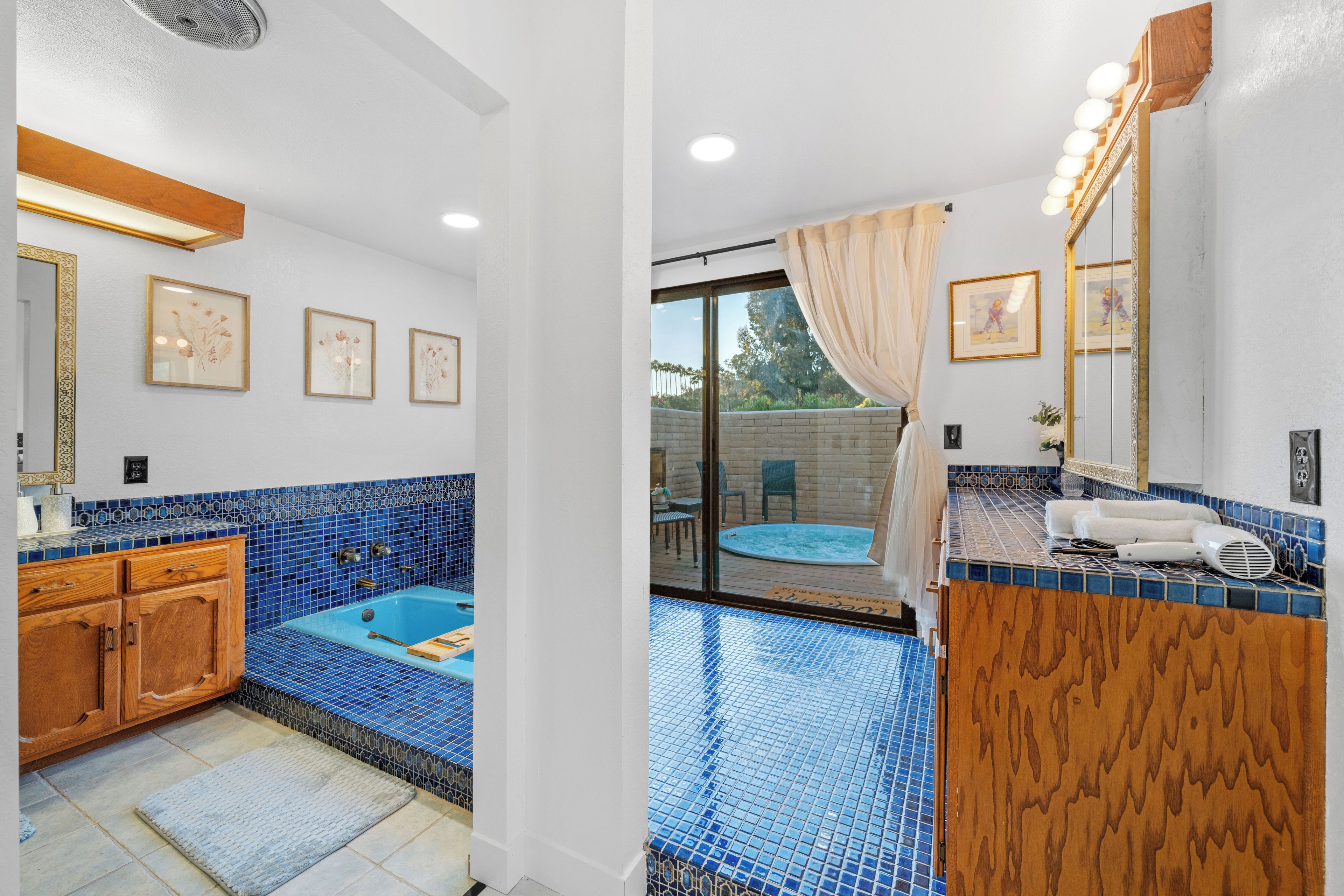 A bathroom with a blue-tiled bathtub, wooden vanity, and a large mirror with lights.