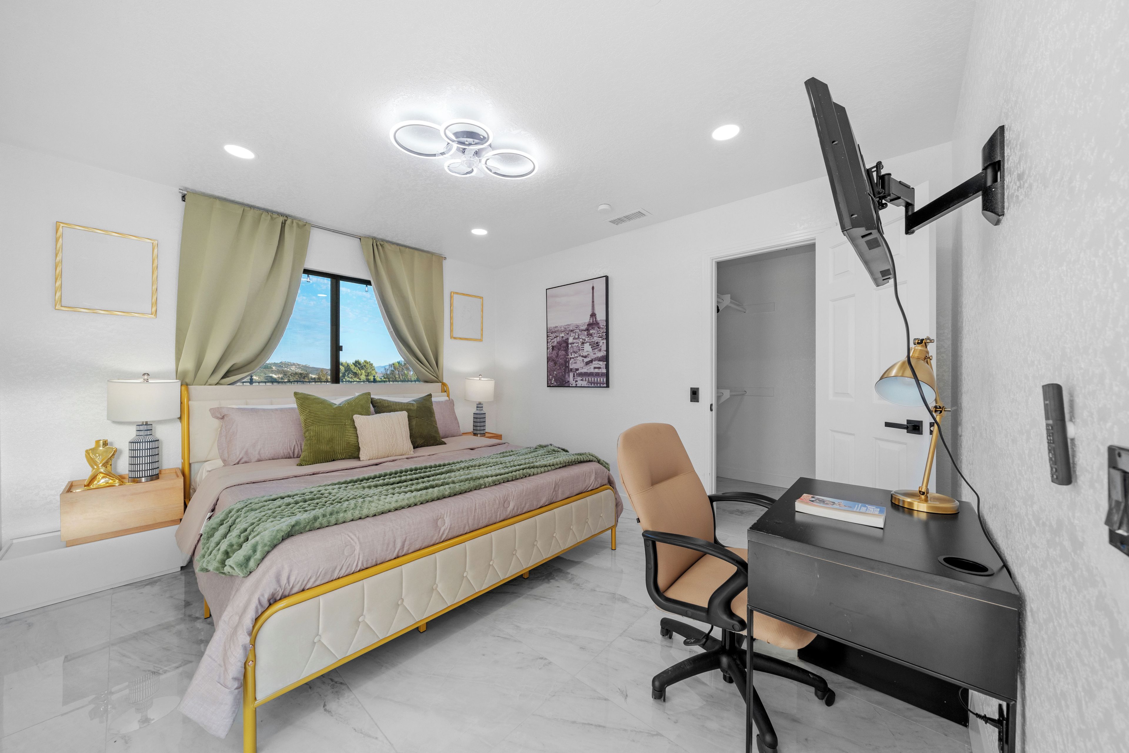 A modern bedroom with a bed, desk, and chair, a flat-screen TV, and a cityscape view.