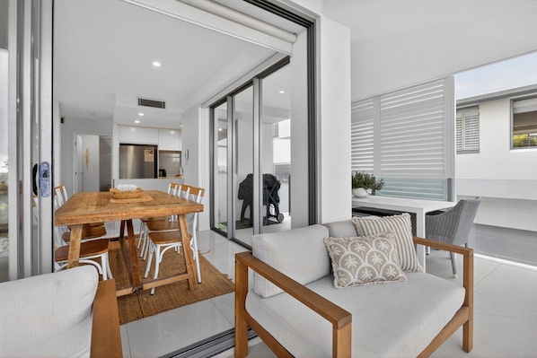 An open-plan living area with a dining table and chairs, a sofa with cushions, and a sliding glass door leading to a balcony. - Soul@Sunshine - Private Pool, Ocean View (Sunshine Beach)