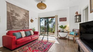 Una sala de estar con un sofá rojo, un tapiz grande en la pared, una lámpara de techo tejida y vistas a través de un ventanal.