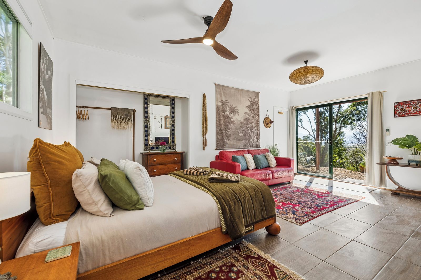 A bedroom with a bed, a red sofa, a ceiling fan, a large window, and a view of the outdoors.