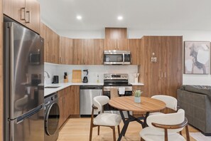 A modern kitchen with stainless steel appliances, wooden cabinets, and a dining area with a round table and chairs. - Elegant Skyline Retreat | 2BR•UG Parking (Edmonton)