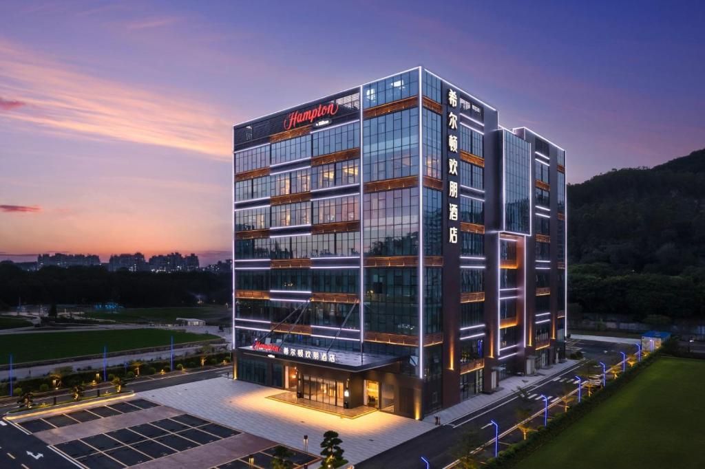 Um hotel moderno de vários andares com uma placa chamativa da 'Hampton', fachada de vidro e sinalização iluminada.