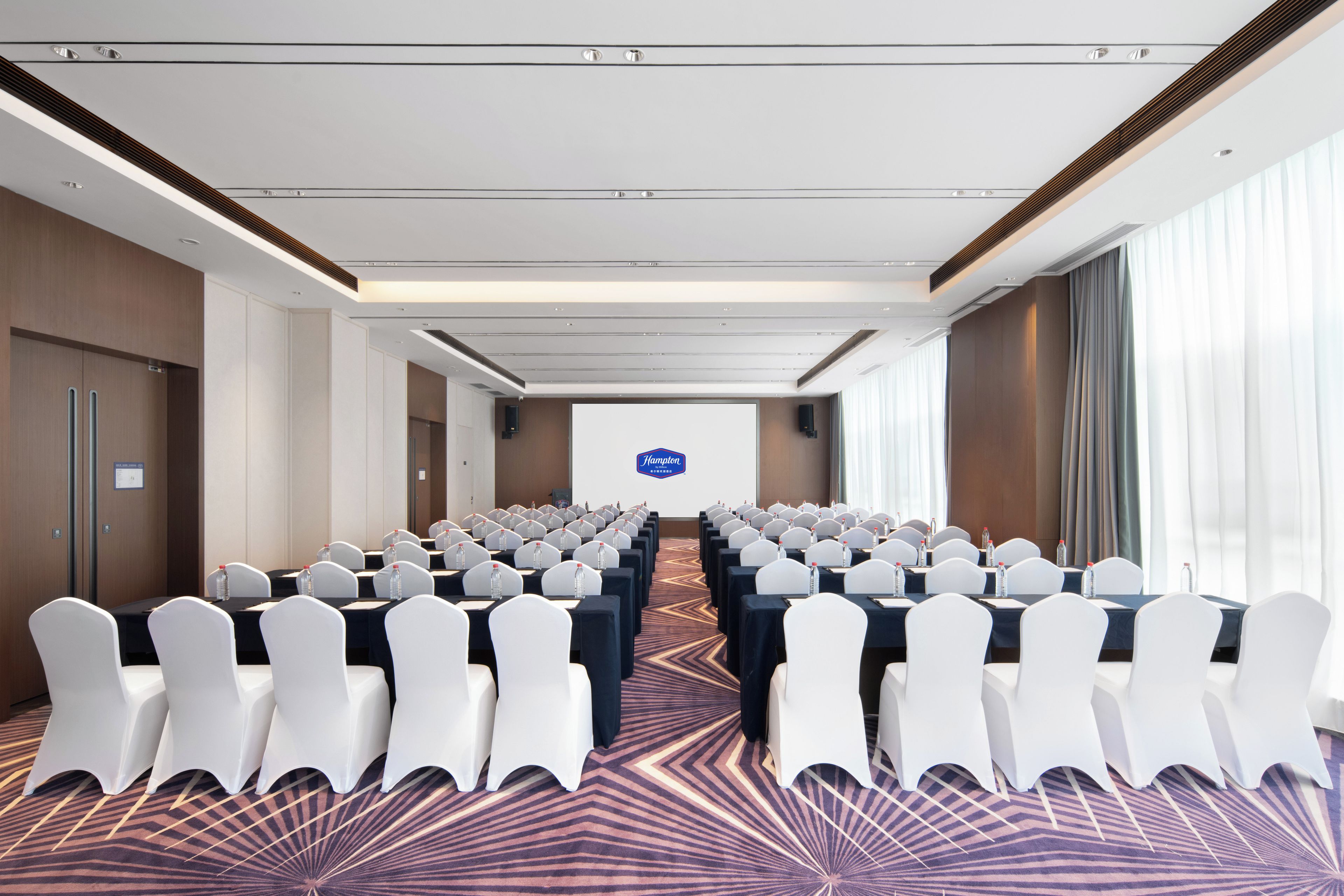 A conference room with rows of tables, white chairs, and a projector screen.