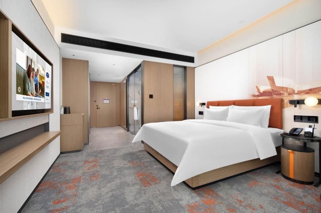A modern hotel room with a large bed, a bedside table with a lamp, a wall-mounted TV, and a glass door leading to a bathroom.