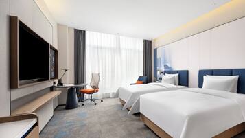 A hotel room with two beds, a desk, a flat-screen TV, and a large window with curtains.