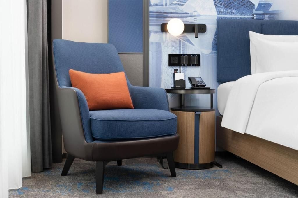 Habitación de hotel con una cama individual, una silla con cojín naranja, una mesita de noche con lámpara y un teléfono instalado en la pared.